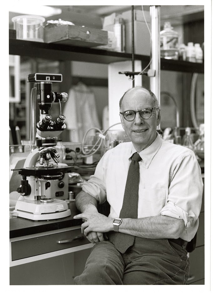 black and white photo of man seated next to microscope