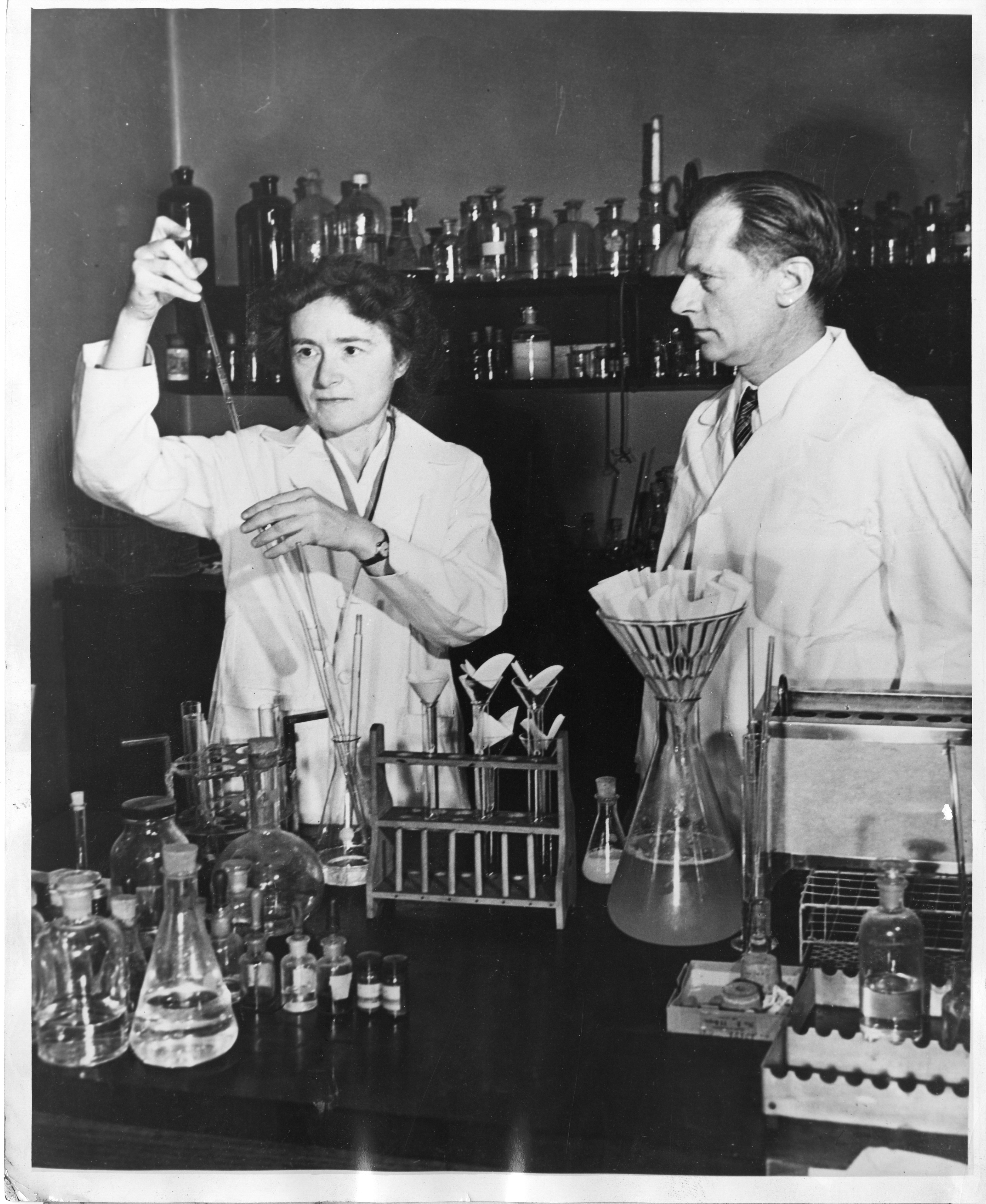 black and white photo of a woman and a man standing in a lab with beakers all around