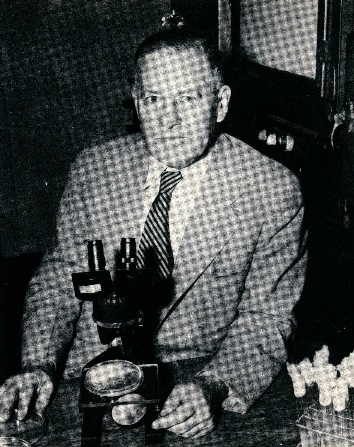 back and white headshot photo of man seated in front of microscope